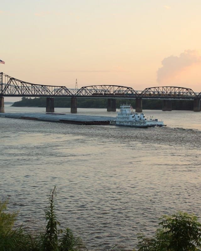 A towboat pushes barges under the Vicksburg bridges at dusk, seen from the bank at MS Riverbend