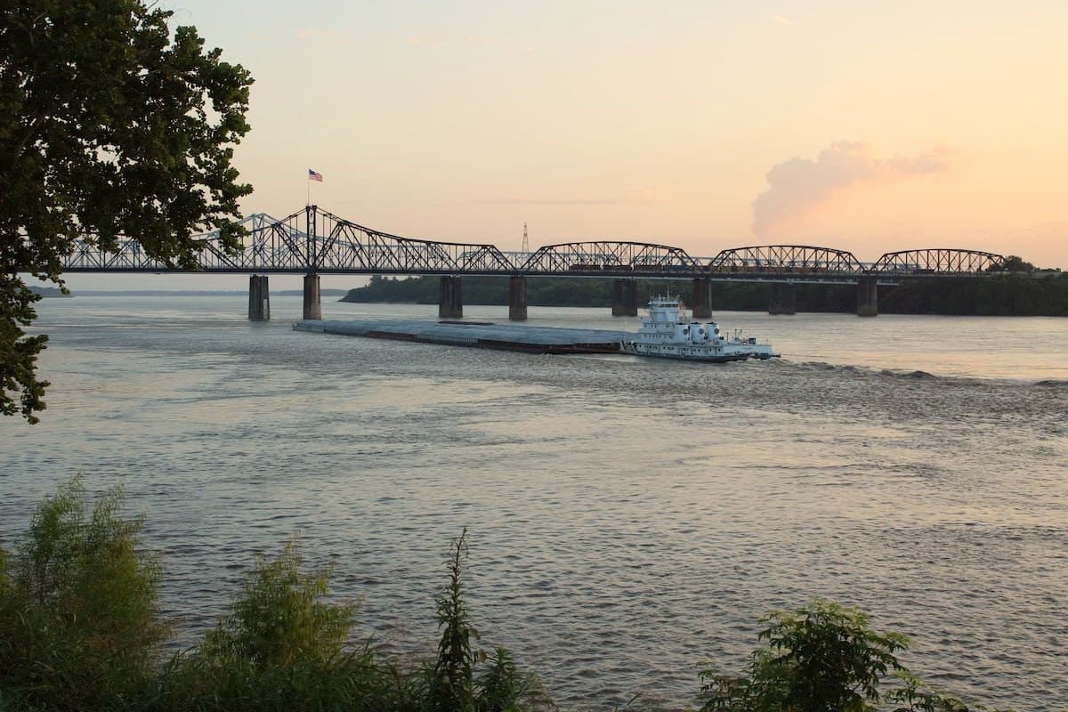 A towboat pushes barges under the Vicksburg bridges at dusk, seen from the bank at MS Riverbend