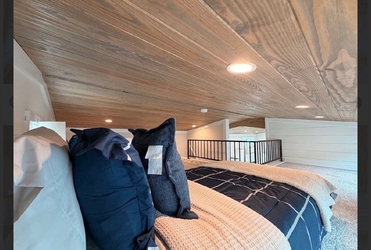 Loft bed with navy pillows under a wood-plank ceiling, stair rail behind