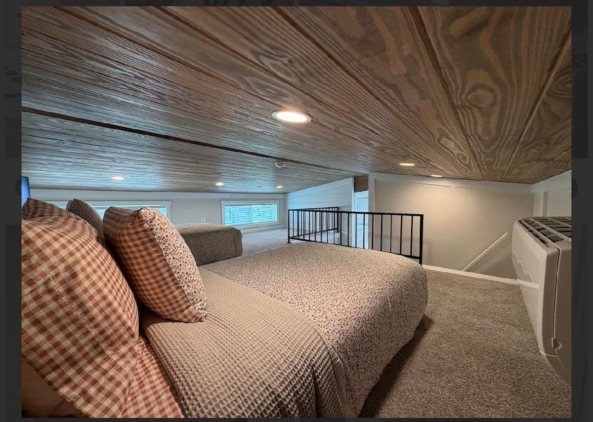 Loft bedroom under a wood-plank ceiling, king bed with tan spread and orange pillows, black stair rail