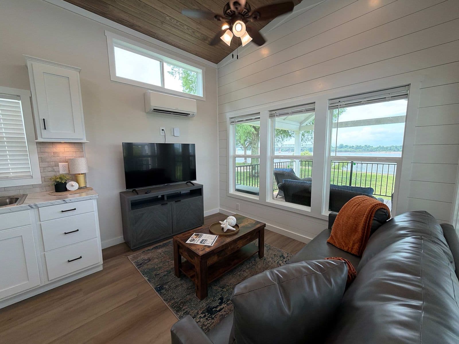 Living area with black leather sofa, TV, orange pillow and river windows