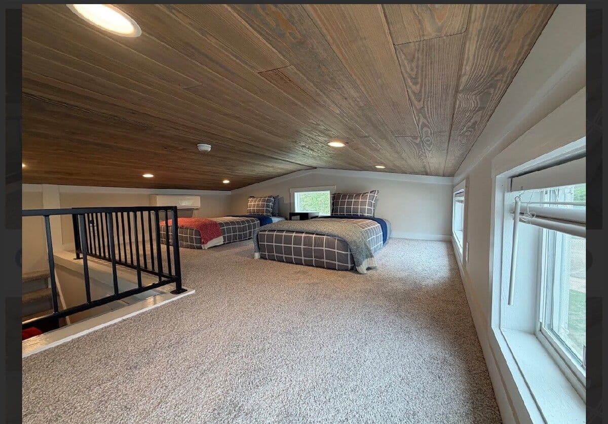 Loft from the stair rail: two beds in plaid spreads under a plank ceiling, big window