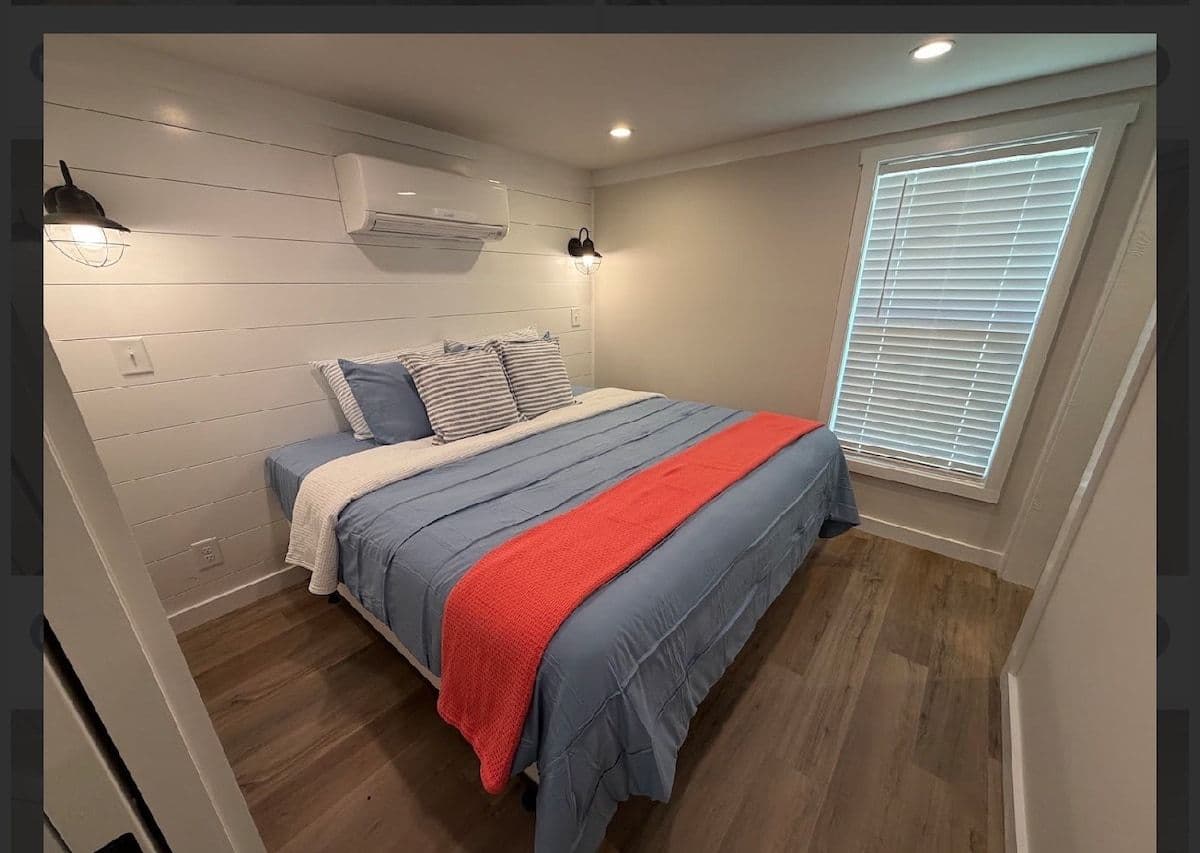 Bedroom with blue spread and red throw, wall AC and window shutters