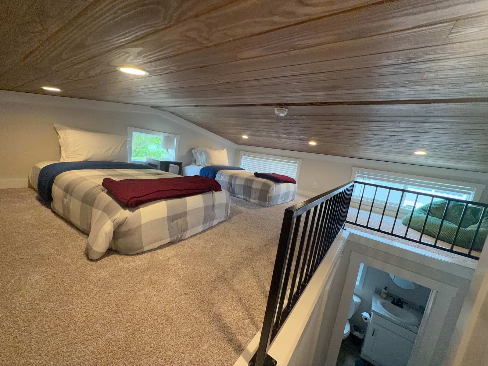 Loft with plaid-spread bed under a wood ceiling, stair rail
