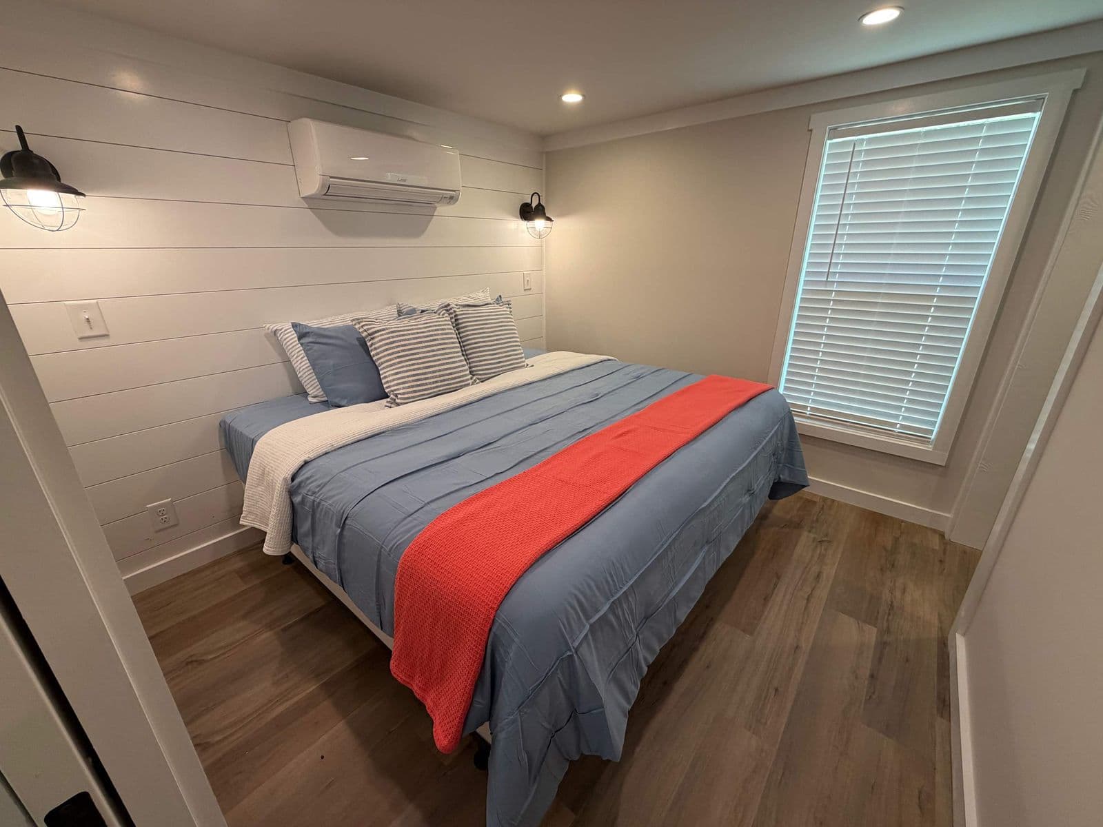 Bedroom with blue spread and red throw, wall AC and shuttered window
